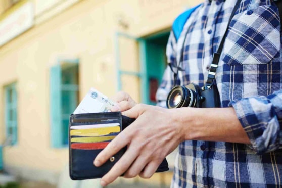 A traveler holds an open wallet with currency and documents, with a camera around their neck, standing outdoors in front of a yellow building, highlighting global travel insurance services.