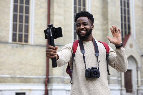Young man smiling, holding a selfie stick with a phone, wearing a backpack and a camera around his neck, standing in front of a historic building.