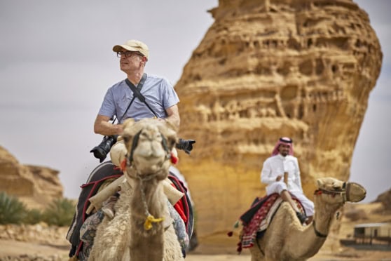 A senior man with binoculars and a camera rides a camel in a desert landscape with large rock formations, highlighting international travel and adventure tourism.