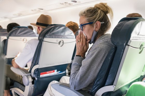 A woman on a plane sneezing into a tissue, wearing glasses and a mask, surrounded by other passengers, illustrating travel health protection for global travelers.