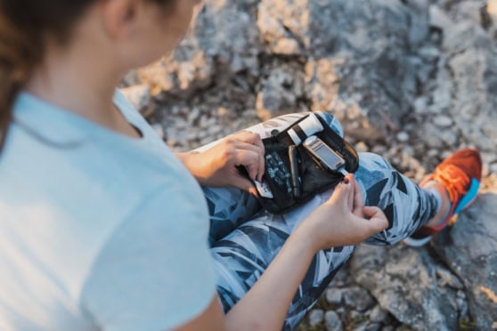 A young woman outdoors on a rocky trail, opening her backpack to access an emergency kit with first aid supplies and essential tools, highlighting travel safety and preparedness.