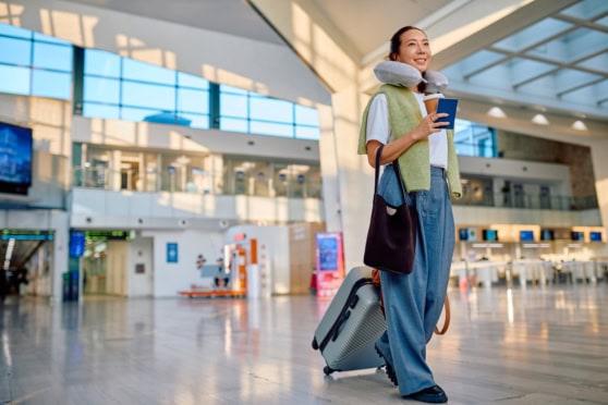 A smiling woman with neck pillow, travel bag, and suitcase waiting in an airport terminal, showcasing international travel preparedness and comfort for global citizens.