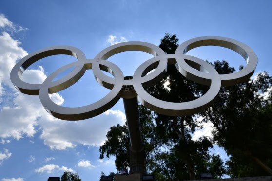 An iconic Olympic Rings monument featuring five interlocking rings symbolizing unity and sportsmanship, set outdoors with natural sunlight and trees in the background.