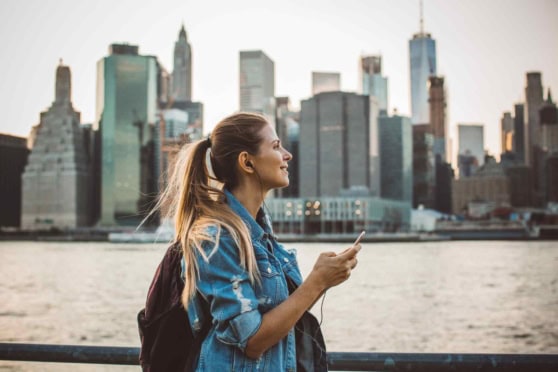A smiling woman in a denim jacket stands by the water with NYC skyline, promoting global health and travel insurance for international citizens.