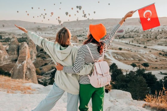 Alt text: Two women in casual clothing overlooking a scenic landscape of Turkey with hot air balloons, one holding a Turkish flag, highlighting travel and international insurance.