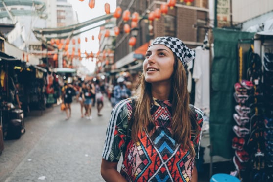 Young woman exploring vibrant street market, wearing colorful attire and patterned headscarf, with shopping stalls, lanterns, and bustling crowd in background.