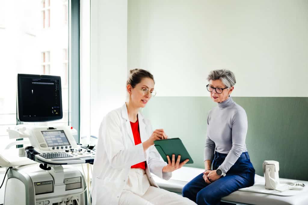 Doctor advising senior woman patient in hospital, showcasing insurance coverage options via tablet, surrounded by medical devices, promoting international health insurance services.