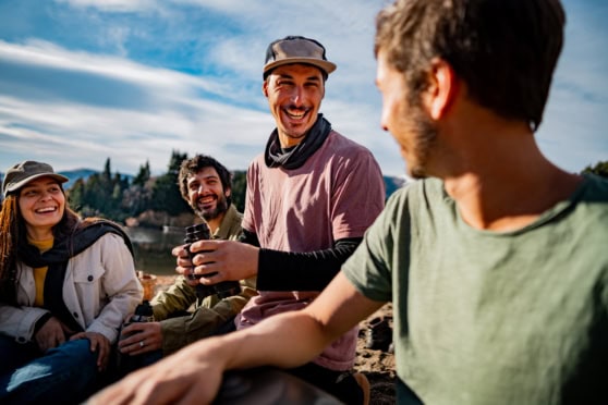 Happy group of friends smiling and sharing moments outdoors near a lake under a blue sky.