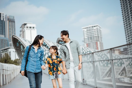 Family of three on a city walk smiling, holding hands, with buildings and a modern urban background.