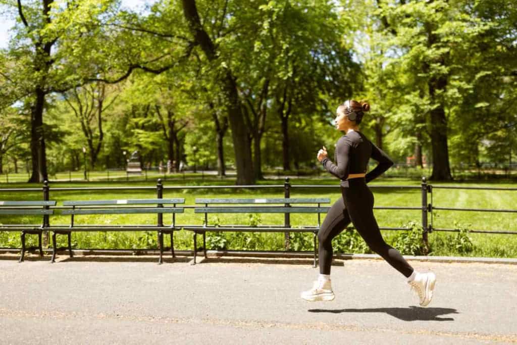 A woman jogging in a lush green park with tall trees and a paved pathway, enjoying outdoor exercise on a sunny day.