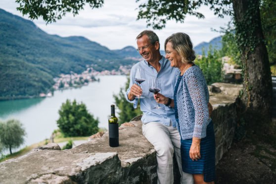 Smiling elderly couple drinking red wine outdoors with a mountain and lake backdrop, emphasizing health, leisure, and lifestyle for international travelers or retirees.