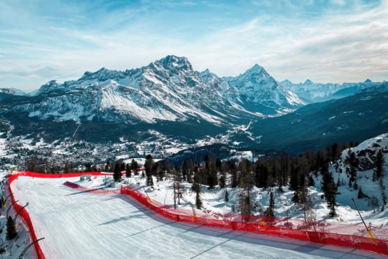 A scenic winter landscape showing a snow-covered ski run with a backdrop of towering, snow-capped mountains and a bright, partly cloudy sky.