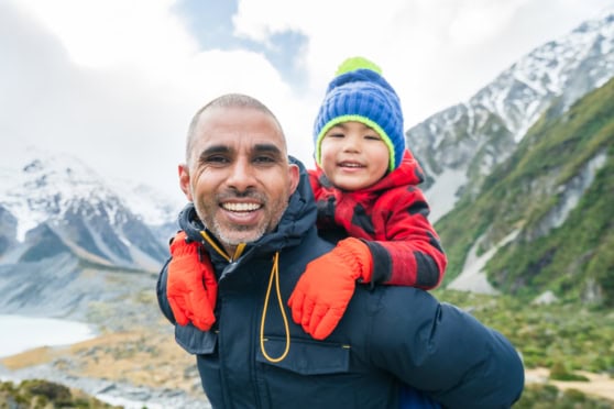 A joyful father carrying his young child on his back during an outdoor mountain hike, set against a backdrop of snow-capped peaks and lush green slopes, embodying adventure, family bonding, and travel safety.