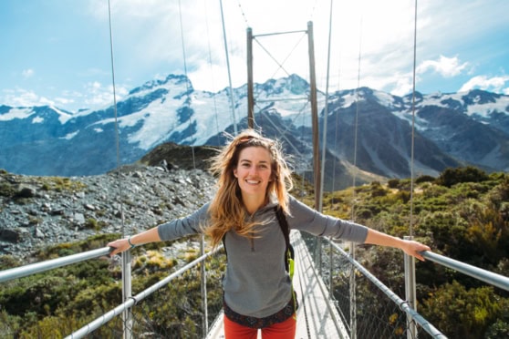 A smiling woman standing on a suspension bridge in snow-capped mountains, illustrating the importance of travel insurance for international travelers and adventurers.