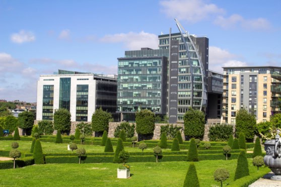 A vibrant cityscape featuring sleek, modern office buildings with extensive glass windows and a beautifully maintained garden in the foreground, highlighting urban living and real estate appeal.