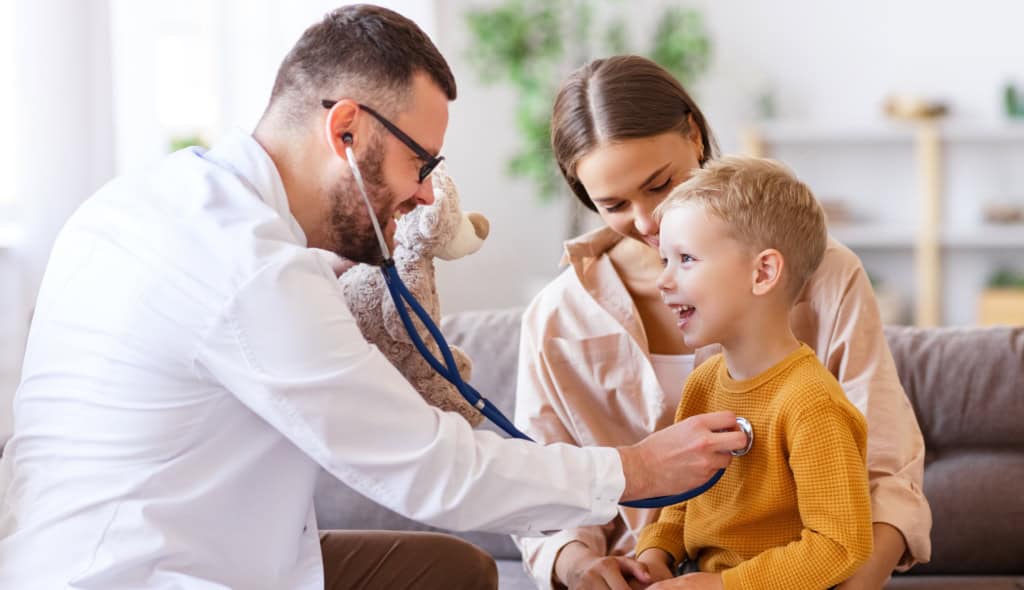 Friendly doctor checking a young boy’s heart with a stethoscope while his mother looks on lovingly in a cozy, well-lit living room.