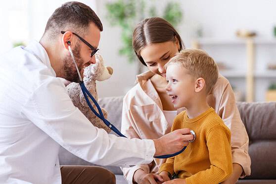 Friendly doctor checking a young boy’s heart with a stethoscope while his mother looks on lovingly in a cozy, well-lit living room.
