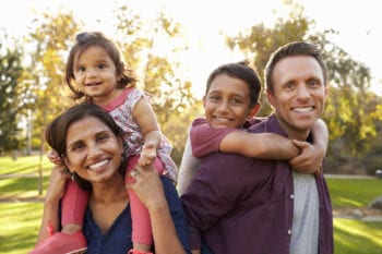 Mixed race parents carry their kids piggyback in a park Mixed race parents carry their kids piggyback in a park
