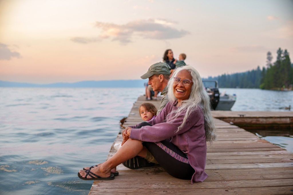 Happy multigenerational family sitting by the lake at sunset, relaxing on a wooden dock, enjoying the peaceful waterside scenery and quality time together.