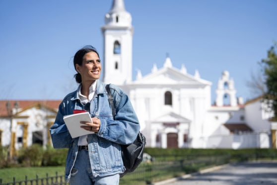 A smiling woman tourist with a notebook and backpack touring historic white church in sunny weather.