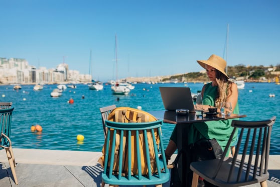 Woman in a green dress and wide-brimmed hat working on a laptop by the harbor in a sunny coastal city.