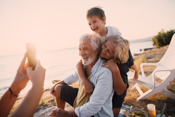 Smiling multigenerational family on the beach at sunset, taking a selfie while kids and grandparents enjoy outdoor leisure time.
