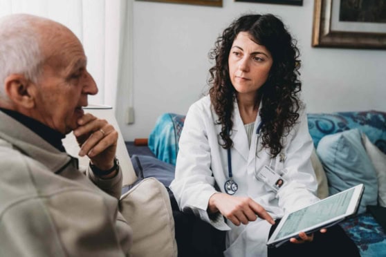 A healthcare professional discusses medical information with an elderly patient in a home setting, using a tablet device to review health details, emphasizing personalized care and international health coverage options.