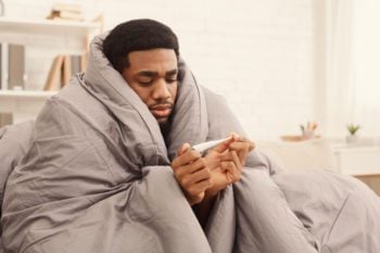 man bundled up in blanket looking at thermometer, who is sick while abroad