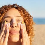 a smiling happy woman applying sunscreen to her face on the beach, having read our expert skin care tips for living abroad