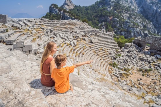 Spectators sitting on stone seats in an ancient Greek theater, surrounded by mountainous landscape. Ideal for those seeking global health coverage and travel insurance options while exploring historical sites abroad.