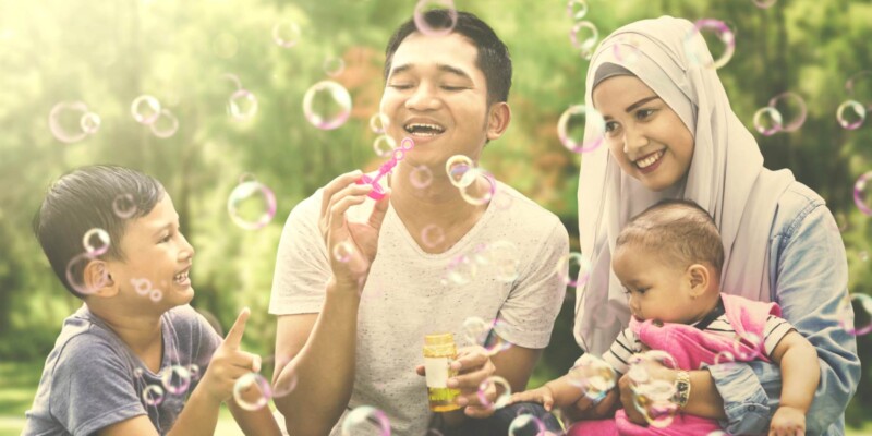 A happy Indonesian family blowing bubbles outside, feeling safe knowing they are protected by insurance for Indonesian citizens