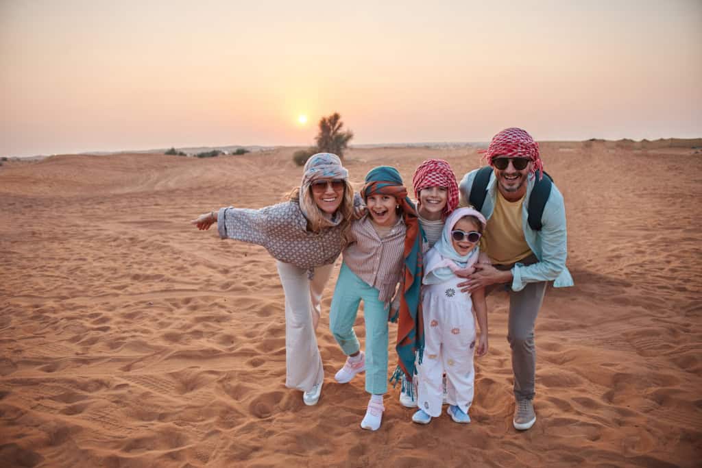 A cheerful family of five dressed in casual clothing and headscarves posing in the desert at sunset, symbolizing travel safety and international adventures.