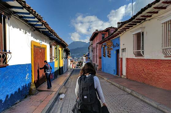 A woman with a backpack walking down a cobblestone street in a picturesque Latin American town, showcasing colorful buildings and scenic mountain views.