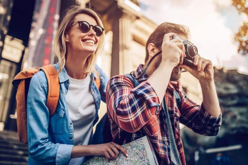 Photograph of a smiling woman and man street photography, woman with sunglasses and backpack, man taking photos with camera, cityscape background, travel adventure.