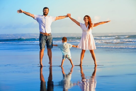 Family at the beach, mother, father, and child holding hands, smiling, with ocean background, emphasizing international health coverage for travelers and expatriates.