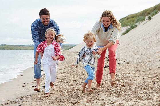 Family smiling and running on the beach, children and parents having fun together in a scenic coastal setting with dunes and water in the background.