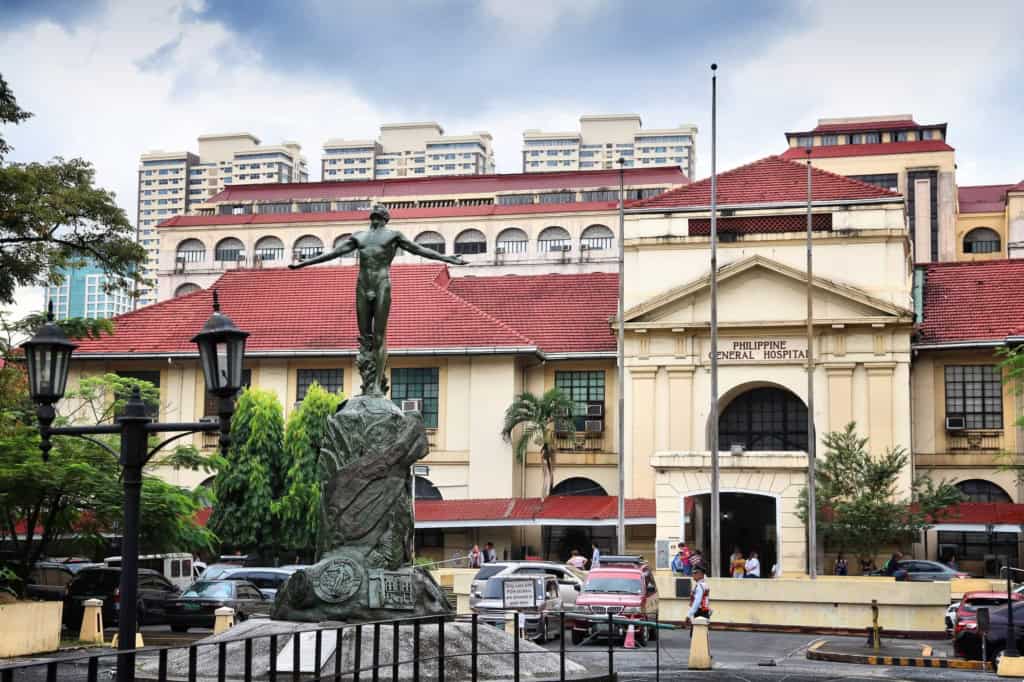 A historic hospital building in the Philippines with a statue in front, surrounded by trees and parked cars, highlighting healthcare coverage options for international citizens.