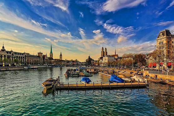 Colorful boats moored at a harbor along the calm river with city architecture and a beautiful sky in the background.