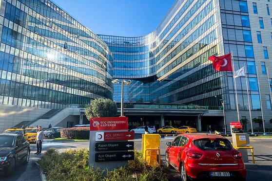 An image of a contemporary hospital with reflective glass windows, Turkish flags, and parking facilities, representing healthcare services in Turkey.