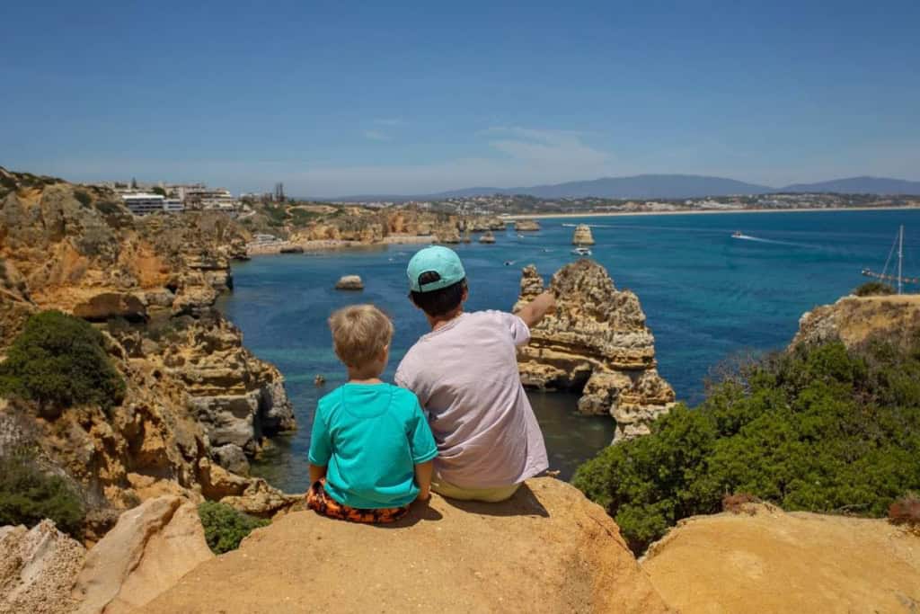 Two children sitting on a rocky cliff overlooking the ocean, enjoying the scenic coastline and clear blue sky. Perfect moment of bonding and exploring nature's beauty.