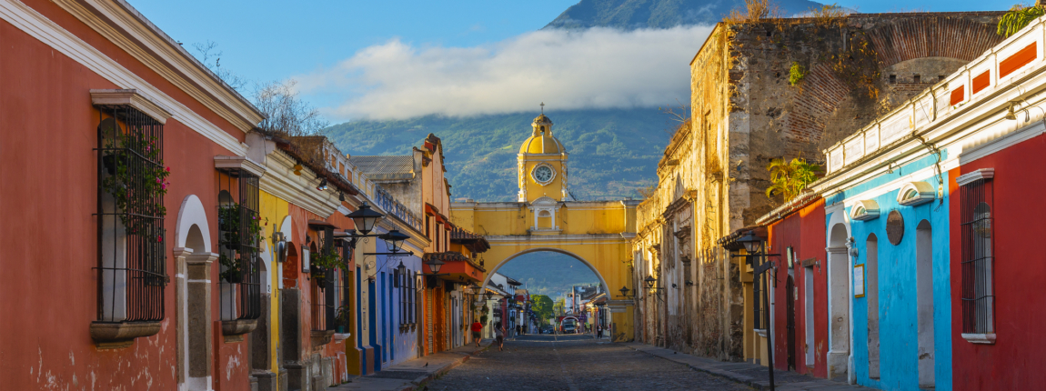 Colorful colonial street in Antigua Guatemala with vibrant buildings and cobblestone pavement, La Merced Arch, and volcano in background, during sunrise.