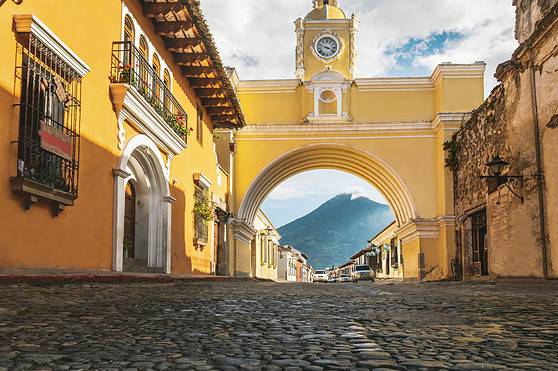 Bright yellow buildings and a historic clock tower in Antigua, Guatemala, with cobblestone streets and a scenic mountain background, ideal for promoting international citizen insurance plans.