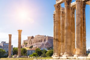 Ancient Greek temple ruins with Corinthian columns and the Acropolis in Athens under bright sunlight, showcasing historic architecture and archaeological significance for tourism and cultural heritage.