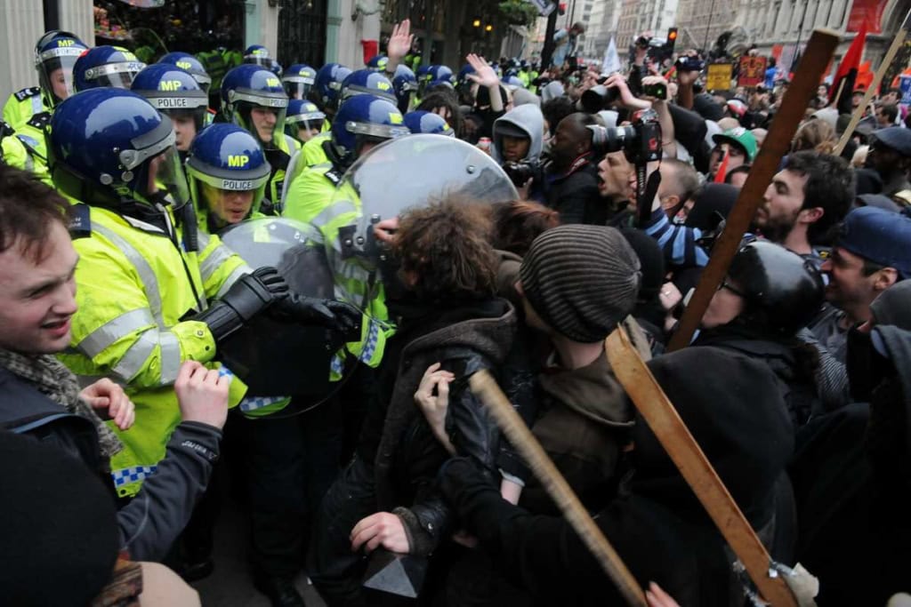 A large crowd of protesters confront police in high-visibility gear, with some holding wooden sticks and shields, on a city street during a tense protest.