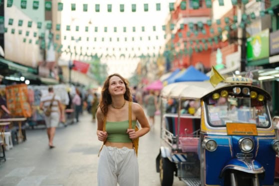 Young woman exploring a bustling street market abroad, smiling and enjoying her trip with an auto rickshaw nearby.