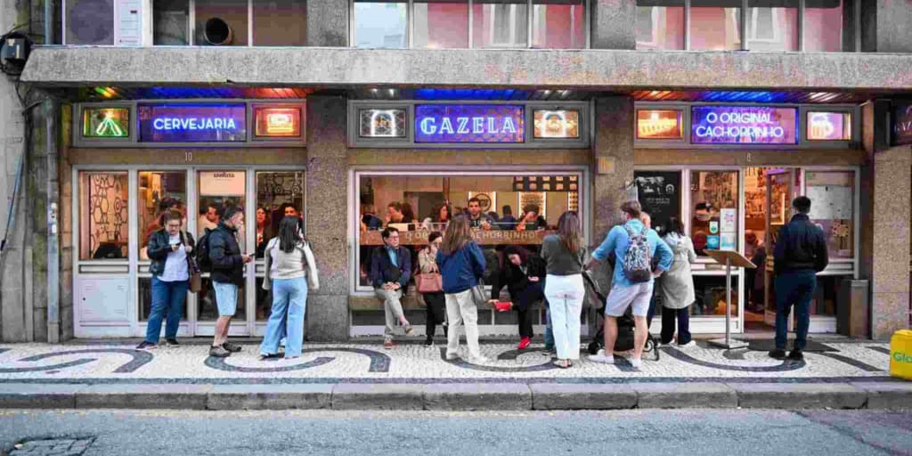 People stand outside a bakery with neon signs reading "Cervejaria," "Gazela," and "O Original Cachorro." Several customers wait in line or chat, enjoying the lively street atmosphere near the bakery.