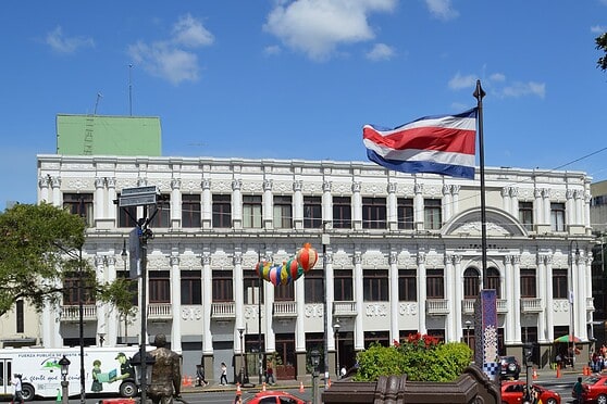 A historic white government building in Costa Rica with a Costa Rican flag waving prominently in front, under a bright blue sky, representing stability and national pride.