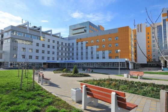 A large, contemporary hospital with white and orange facade, multiple windows, and a welcoming outdoor area with benches and green landscaping under a bright blue sky.