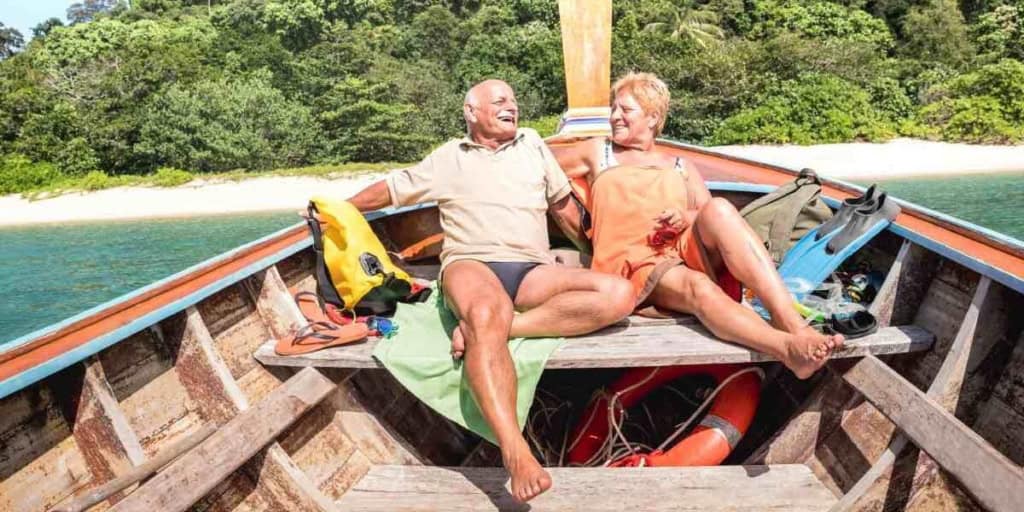 Elderly couple relaxing on a boat deck surrounded by clear water and lush green trees, enjoying a tropical vacation with smiling faces, bright sunlight, and outdoor adventure.