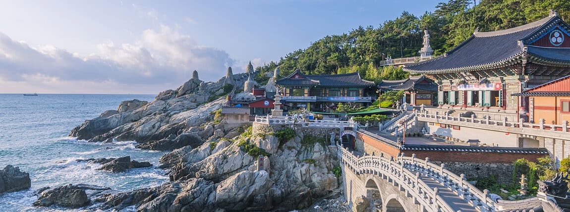 Korea-temple-by-the-sea-on-rocky-coastline-at-dusk, Traditional Korean temple architecture along the scenic coastline with ocean views and lush hillside, ideal for cultural tourism and spiritual retreats, SEO for Korea tourism and cultural heritage.